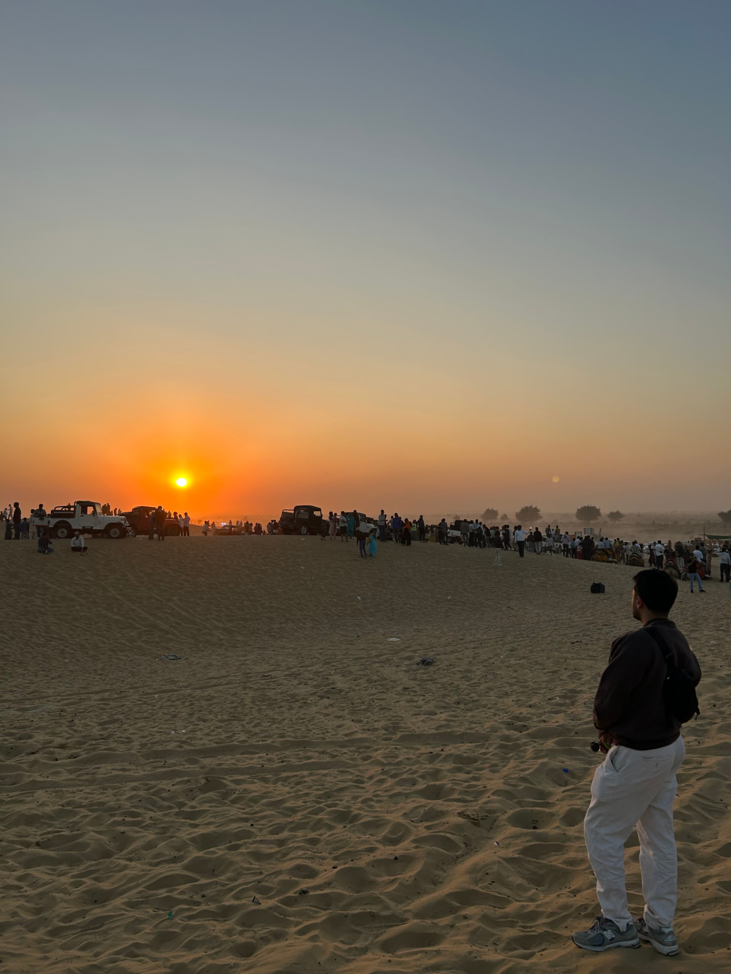 Sunset over sand dunes with jeeps and a crowd gathered on the horizon